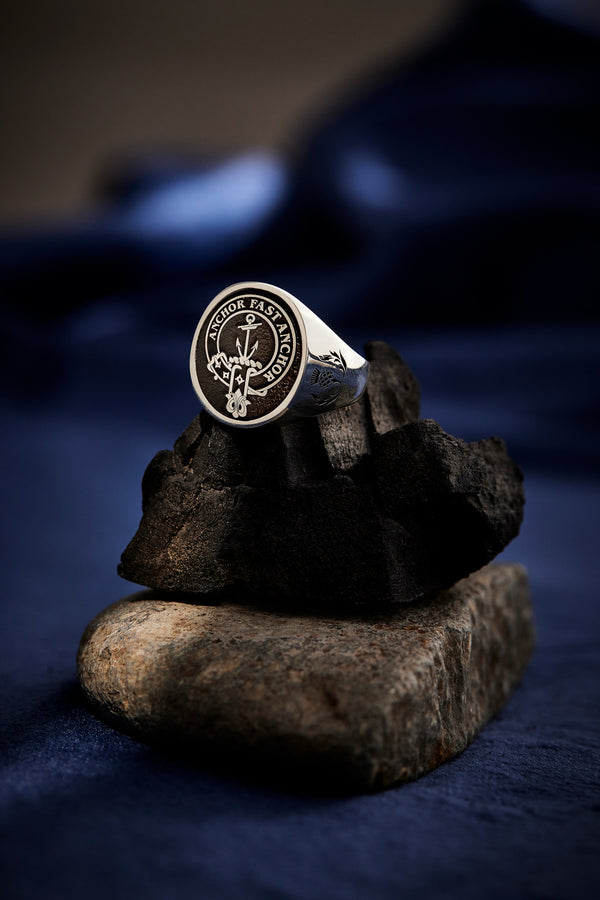 A silver Clan Crest ring engraved with an anchor design and the phrase "Anchor Fast Anchor," displayed on dark stones.