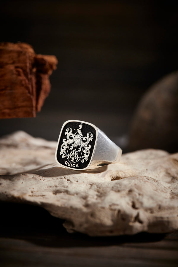 Square silver ring with Quick coat of arms engraved, placed on textured beige rock with dark background.