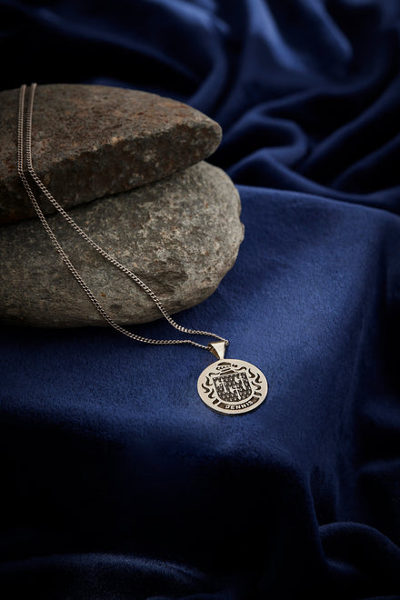 A sterling silver pendant with an engraved crest resting on smooth stones and blue fabric.