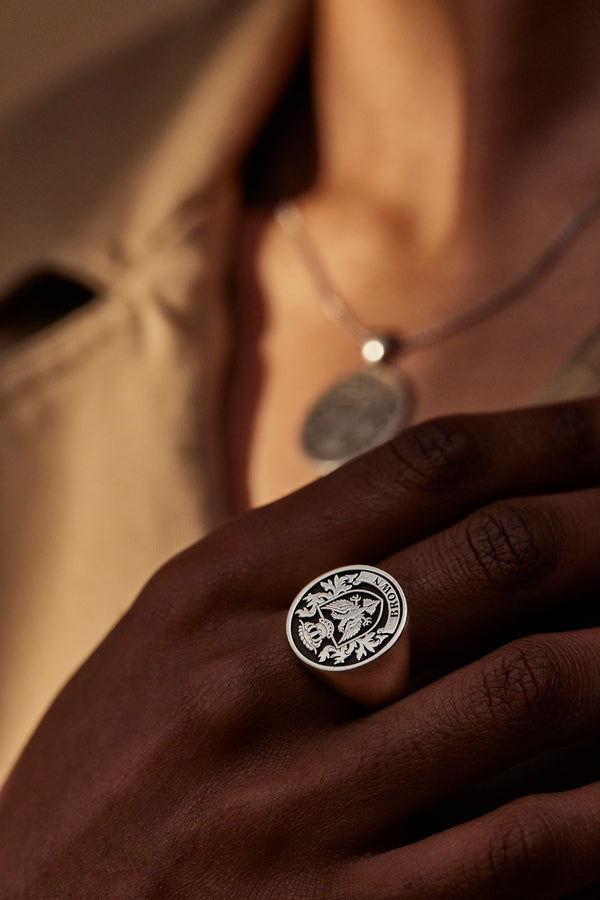 A close-up of a hand wearing a signet ring engraved with a family crest, featuring the name "Brown" and a pendant necklace in the background.