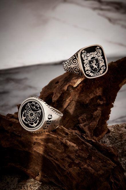 Two sterling silver rings, one with a family crest and the other with a custom crest resting on driftwood.