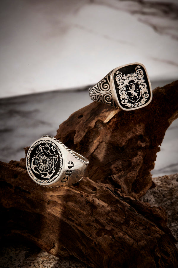 Two intricately engraved rings with coat-of-arms designs, set against a textured, natural wood surface.