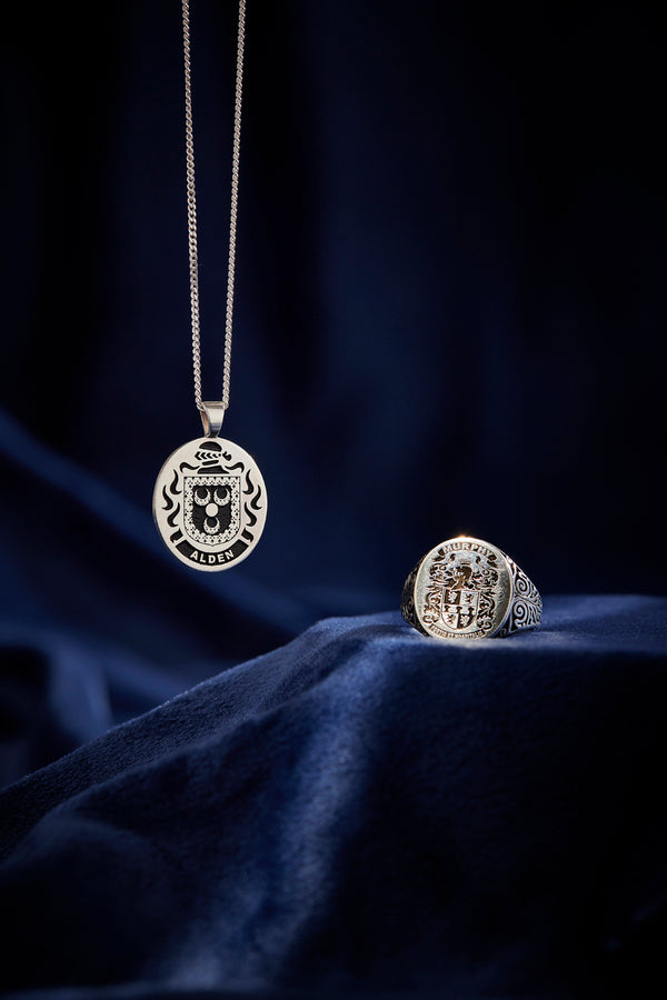 Silver pendant with 'Alden' crest and an intricately designed silver ring with 'Murphy' crest on dark fabric.