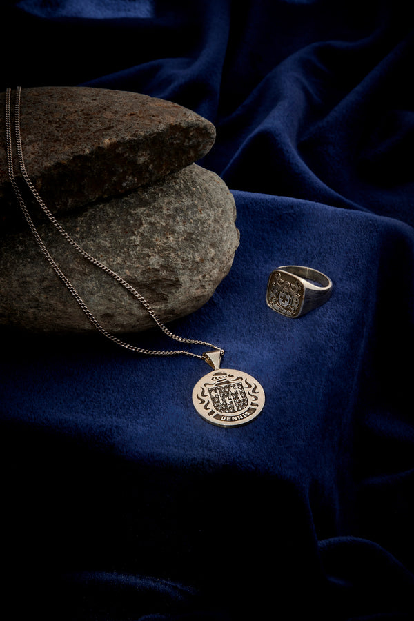 A silver pendant engraved with a coat-of-arms design and the name "Dennis," displayed on deep blue velvet fabric.