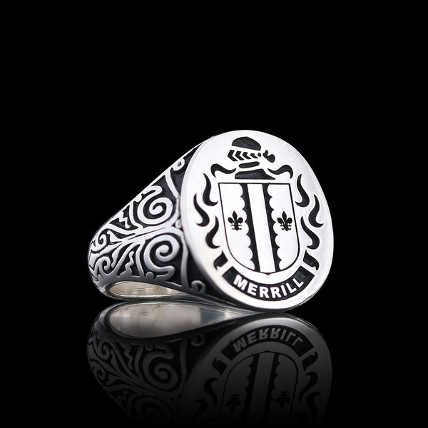 A silver ring with ornate side engravings, displaying the Merrill family crest, including fleur-de-lis and shield details.