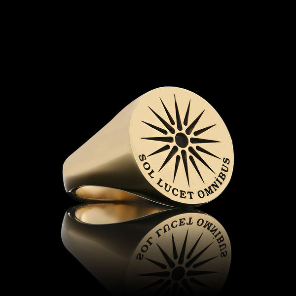 A gold ring engraved with a sunburst design and the Latin phrase "Sol Lucet Omnibus," reflected on a black surface.