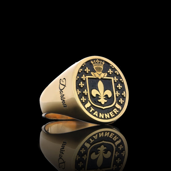 A gold ring engraved with a fleur-de-lis shield design and the name "Tanner," with the name "Dorina" on the band.