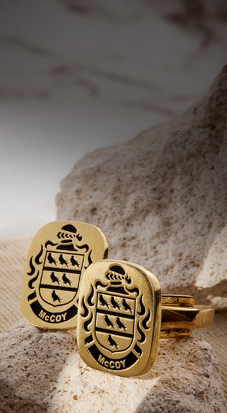 Gold cufflinks featuring the McCoy family crest, placed on textured stone with a neutral fabric background.