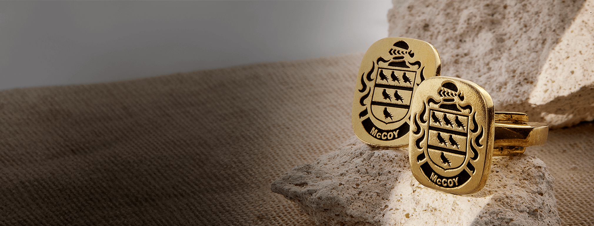 Gold cufflinks featuring the McCoy family crest, placed on textured stone with a neutral fabric background.