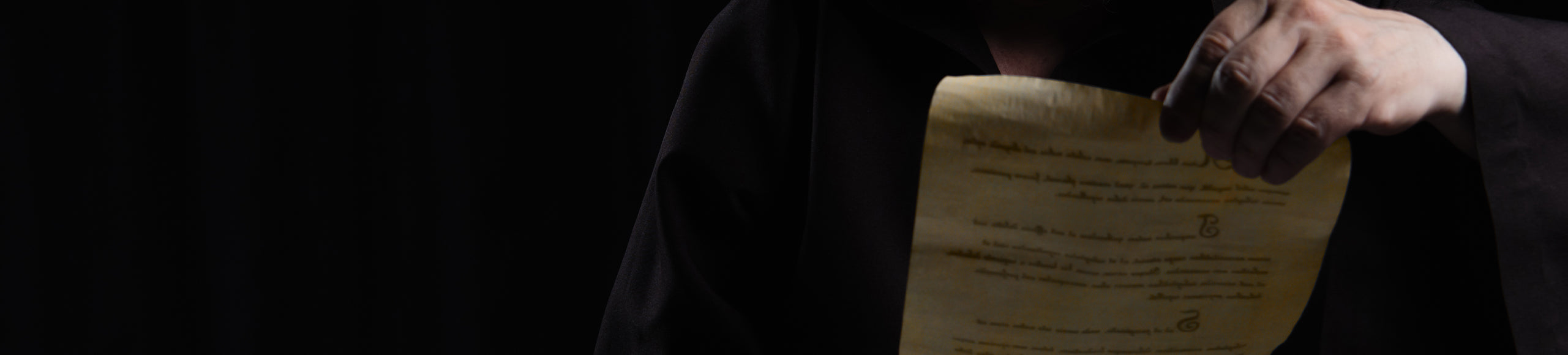 Close-up of a historical document held by a person in dark robes, illuminated under soft lighting.