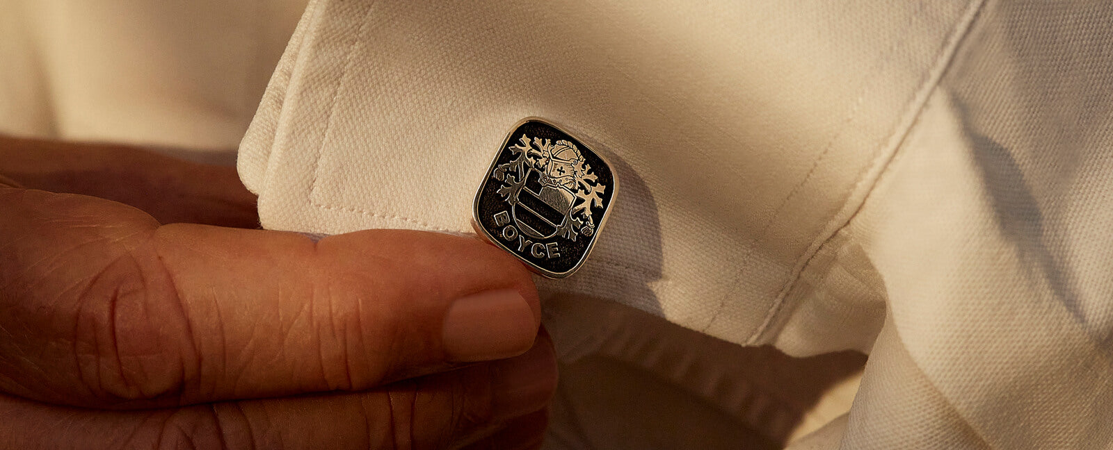 Close-up of a silver cufflink with a family crest engraved, held against the cuff of an elegant white shirt.