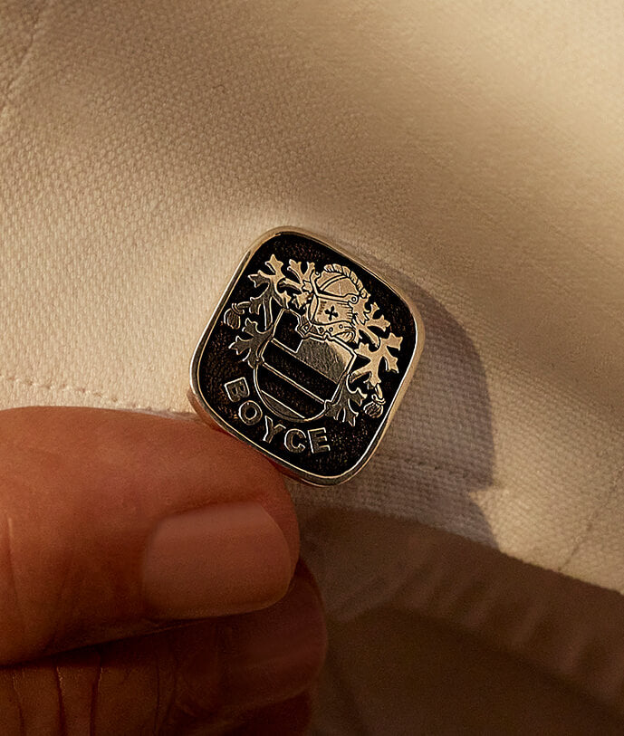 Close-up of a silver cufflink with a family crest engraved, held against the cuff of an elegant white shirt.