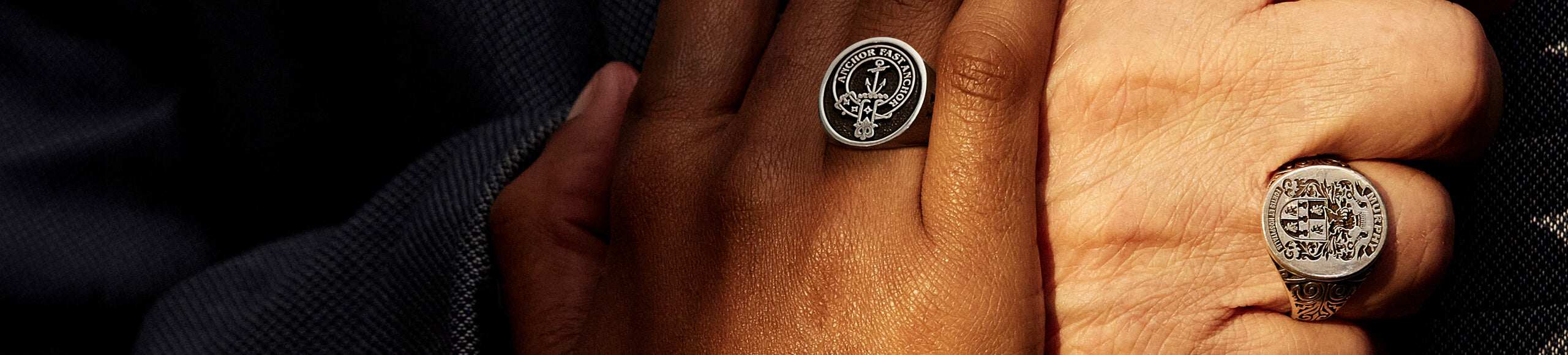 Two hands, one wearing an ornate signet ring and the other a clan crest ring, against a tailored suit.