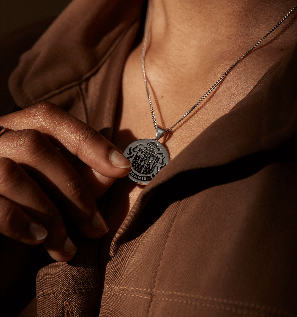 A person wearing a sterling silver crest pendant necklace, gently holding it against a brown outfit.