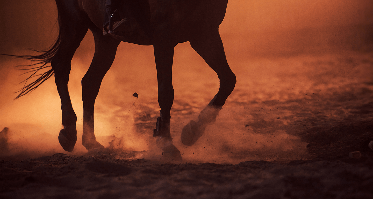 Horse hooves kicking up dust on an orange-lit sandy surface.