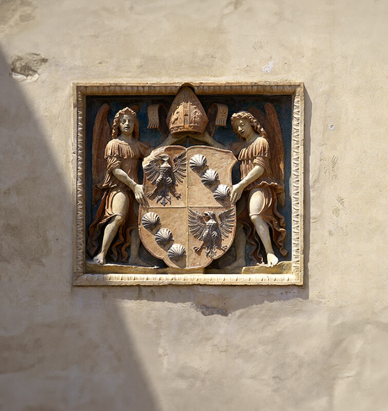 Wall-mounted coat of arms flanked by angelic figures, displayed on a textured beige background, evoking heritage and artistry.