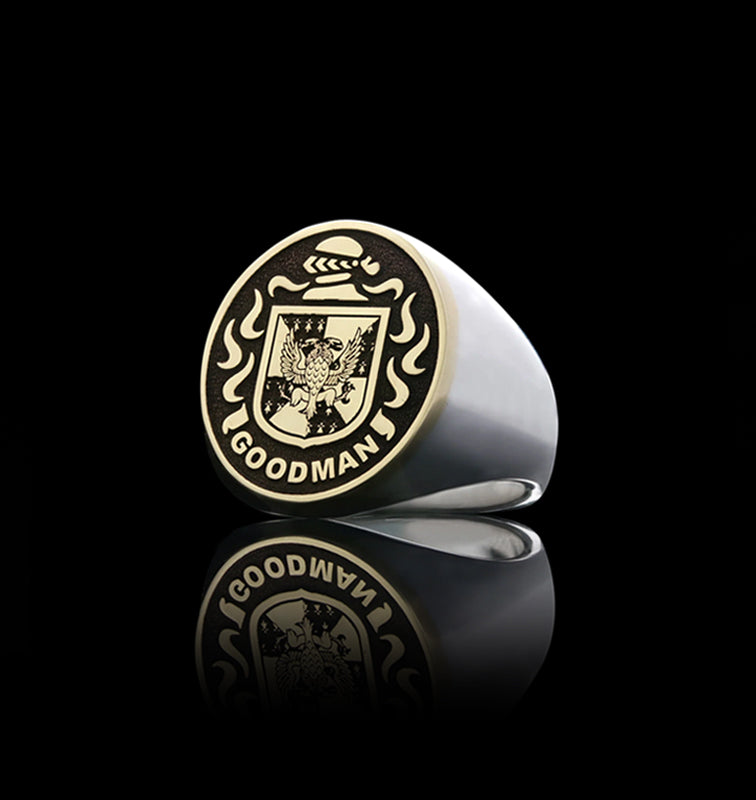 A silver and gold signet ring engraved with the name "Goodman," reflected on a glossy black surface.