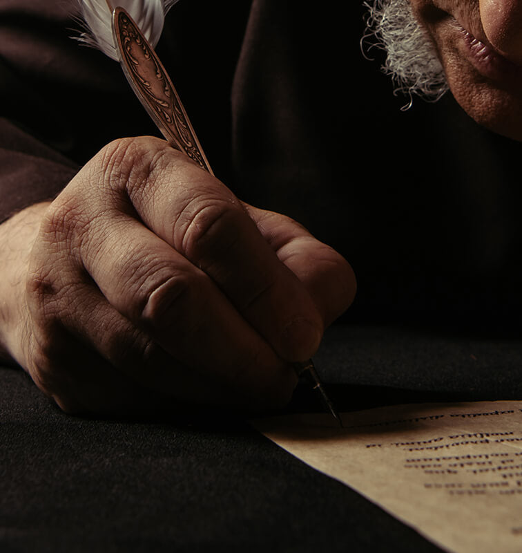 Dark background with a hand writing on parchment using a quill pen.