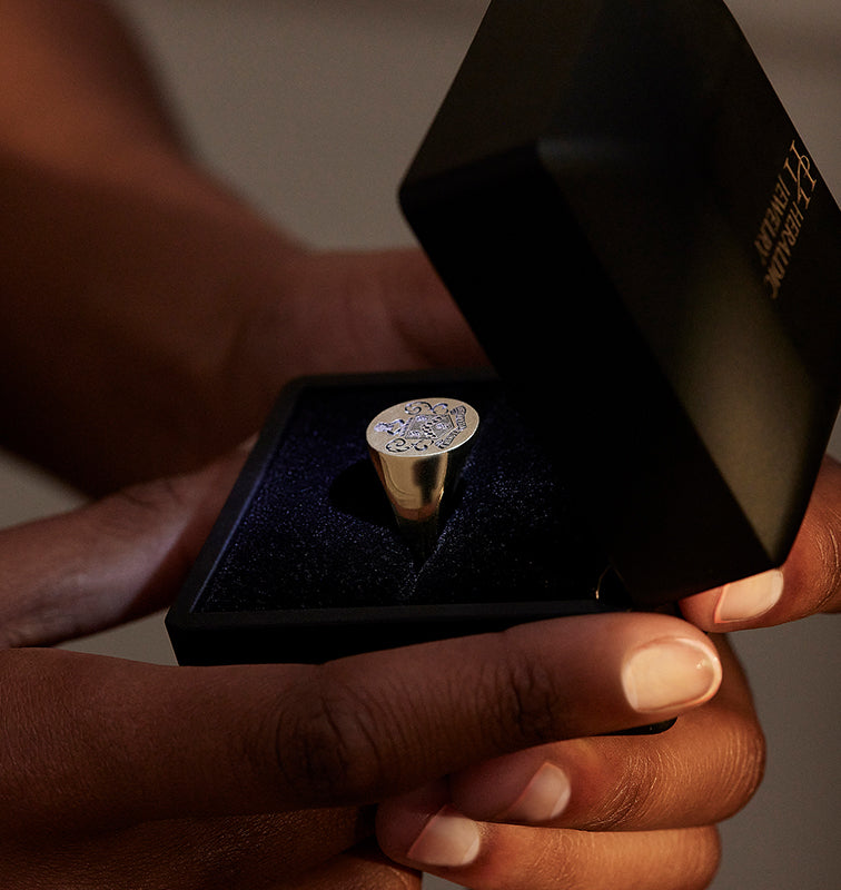 A golden crest ring displayed in an elegant black gift box, held in someone's hands.