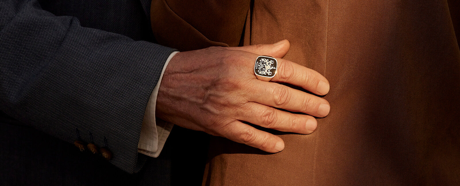 A hand wearing a family crest ring rests gently on a person wearing a brown coat, conveying a sense of heritage and connection.