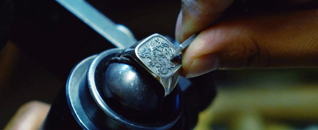 Close-up of a craftsman engraving a detailed family crest onto a silver ring using precision tools.