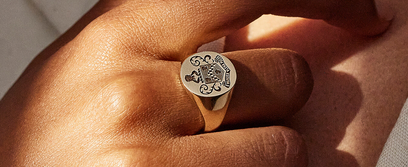 Close-up of a gold signet ring with an engraved family crest worn on a person's finger in natural sunlight.