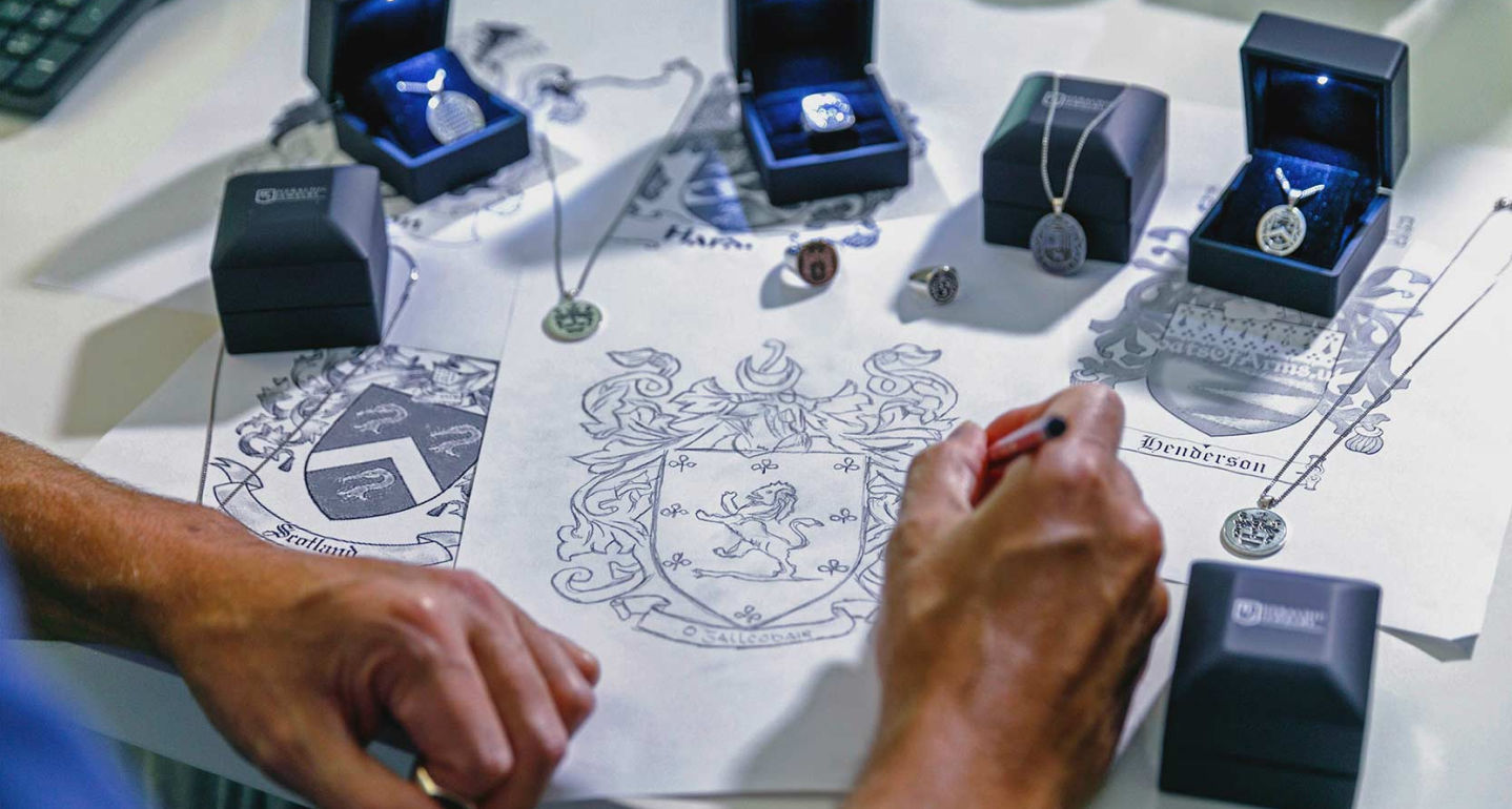  designer sketching a family crest surrounded by rings and pendants in display boxes.