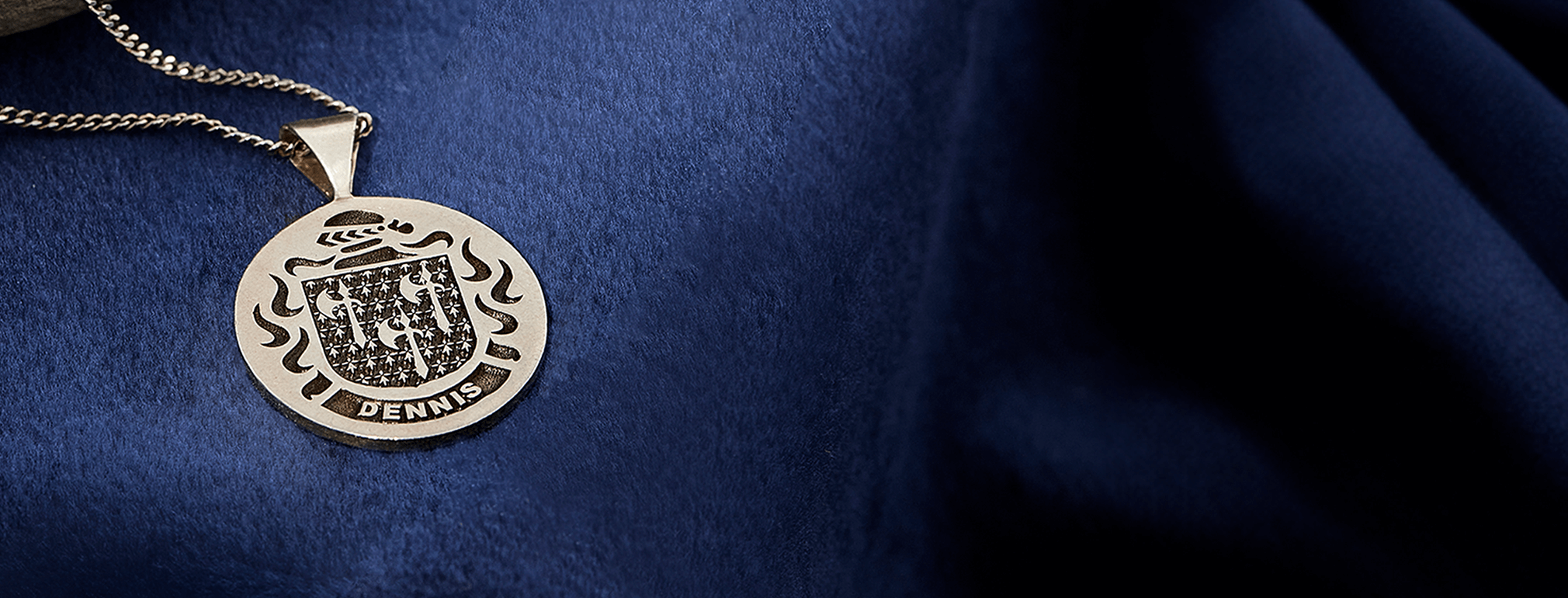 Sterling silver pendant with the Dennis family crest, displayed on a silver chain against a rich blue velvet background.