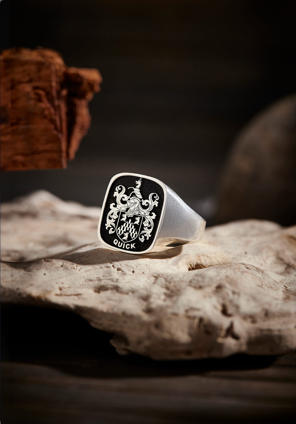 A sterling silver signet ring engraved with the Quick family crest, placed on a weathered stone surface with a wooden backdrop.