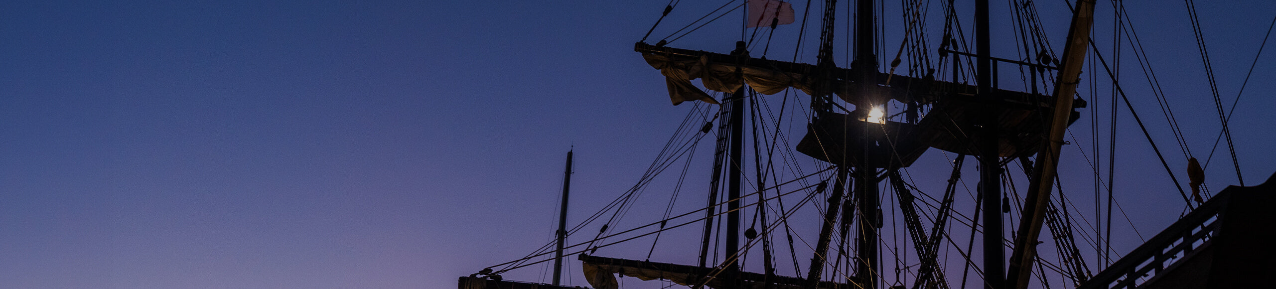 Silhouette of a vintage sailing ship's masts and rigging against a twilight sky.